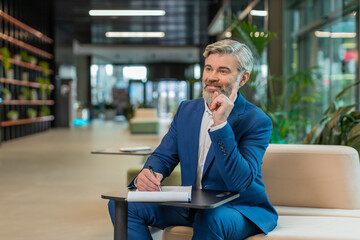 Caucasian middle-aged businessman working with documents, analyzing information, reading papers prepare financial report. Happy male entrepreneur in suit analyzing graph documents in office lobby.
