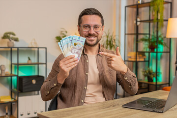 Young businessman freelancer holding fan of cash money Ukrainian hryvnias UAH banknotes, pointing...