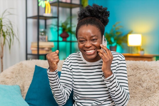 African American woman smiling and gesturing while holding smartphone during a positive phone conversation talk. Enjoying good news relaxed happy. Sitting comfortably at home feeling joyful connected - Powered by Adobe