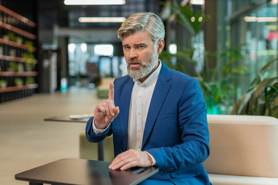 Portrait of mature businessman shakes finger and saying No be careful scolding and giving advice to avoid danger mistake disapproval sign sitting in modern office lobby. Serious guy in suit at hotel.