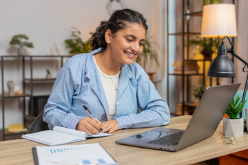 Young woman writing down or taking down notes while attending online video class or office meeting on laptop computer at home office desk. Girl freelancer support services looking at webcam. Lifestyle