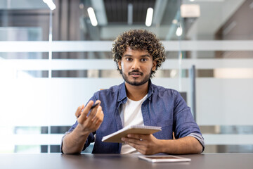 Portrait of a young Indian man sitting in an office in front of the camera, talking online and taking notes in a notebook