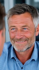 Obraz premium An older Caucasian man with gray hair and blue eyes smiles at another person who is out of view. He wears a blue striped shirt and appears to be engaged in conversation