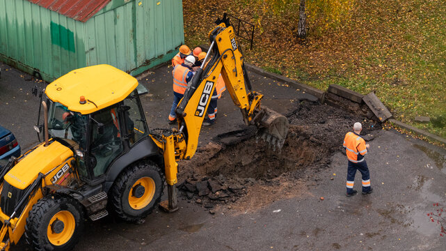 Saint Petersburg, Russia - November 3, 2025: Construction workers in reflective vests and hard hats using yellow excavator to dig trench for street repair.