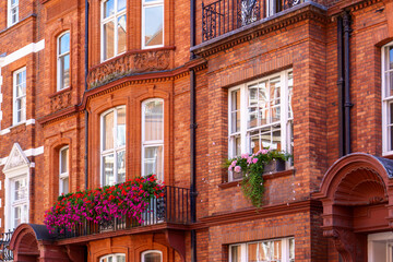 Red brick facade of a British-style building with a small balcony decorated by vibrant flowers.