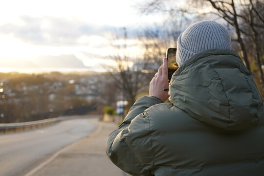 Man taking in panoramic Norwegian sunset with phone, forest below, city ahead,