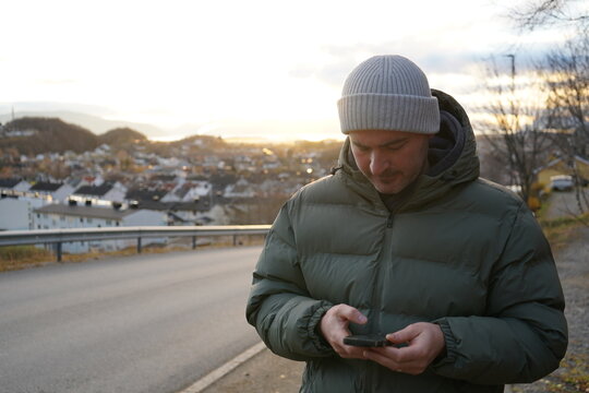 Man scrolling phone above Norwegian city, sunset reflections and warm tones,