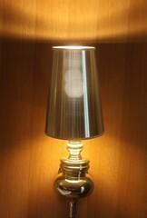 A polished metal table lamp casts a soft amber glow against a wooden wall, creating a cozy, inviting atmosphere. Ideal for bedroom, living room, or hotel decor and lighting staging.