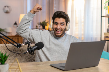 Young Indian man playing online game on laptop actively reacting to game's events during live stream, using headphones, studio microphone while recording vlog sitting at table in living room at home.