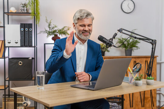 Middle-aged businessman at home office records podcast into studio mic gesturing to highlight ideas. Freelancer guy at table smiles sharing insights engaging audience and refining podcast flow live.