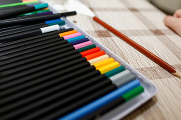 Set of black wooden colored pencils neatly arranged in a case, showing the color tips, next to a single pencil on a checkered table