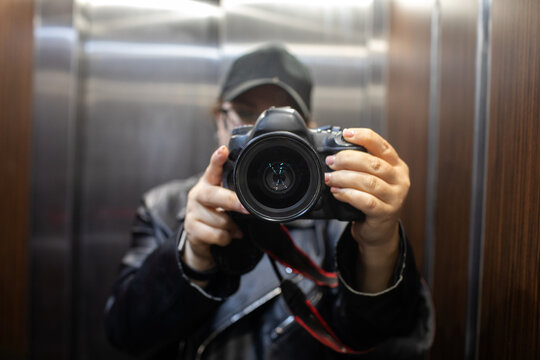 Photographer taking a selfie or video with a dslr camera held up close, standing in a mirrored elevator with stainless steel walls