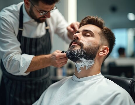 Barber applies shaving foam to man beard with brush. Close up of male client receiving beard trim in salon. Professional grooming service in barbershop.