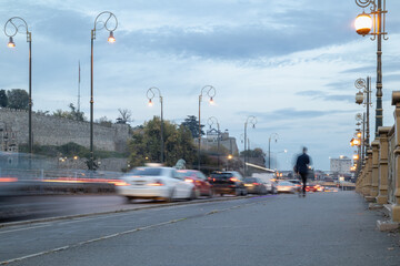 long exposure of evening traffic blurred with light trails on a city road, with historical fortress walls and ornate street lamps under a dramatic sky