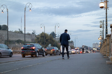 man walking on the pavement of an old bridge at dusk, blurred cars driving slowly in heavy traffic, ancient fortress wall in the background under an overcast sky