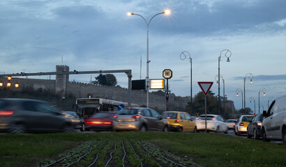 blurred city traffic at dusk in front of an ancient fortress wall and bright street lights, showing movement and urban energy at twilight