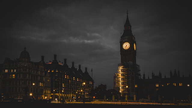 Night view of Big Ben and Westminster under brooding clouds, with warm clock glow and city lights streaking across the frame.