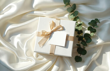 White gift box mockup with blank tag on silky fabric. Ribbon bow and greenery decorates it. Perfect for invitation cards wedding events presents birthdays.