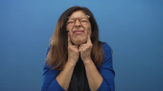 Hispanic middle aged woman wearing glasses and pushing her cheeks with her fingers in studio; playfulness.