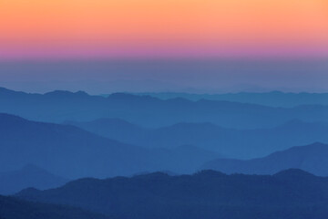 Mountain layers at twilight with colorful gradient sky from orange to blue. Scenic landscape showing misty mountain ridges in silhouette creating depth.Chiamgmai, Thailand. Doi Monjong.