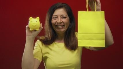 Middle aged hispanic woman wearing yellow shirt holds piggy bank and paper bag against red studio wall; savings planning optimism.