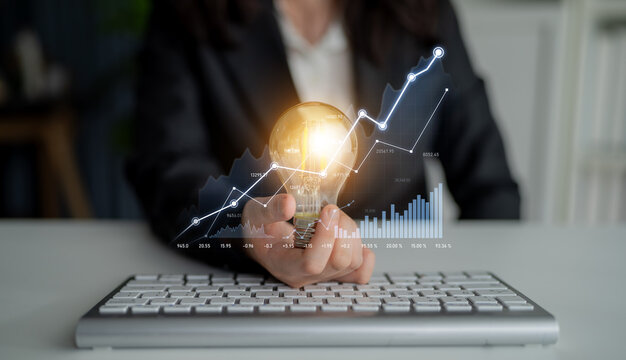 A businesswoman presents a lightbulb while analyzing digital graphs on a keyboard, symbolizing innovation, growth, and strategic planning in the modern workspace. Scalp