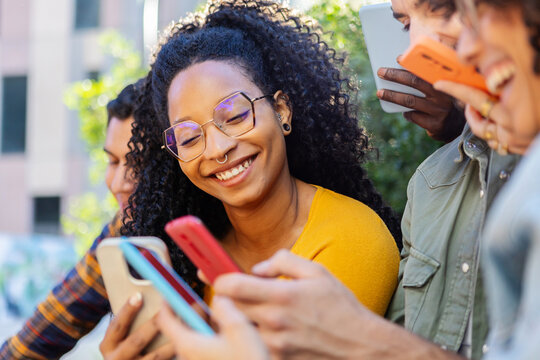 Multiracial young friends enjoying social media on smart phones outdoors. African American woman having fun with young colleagues using cellphones.