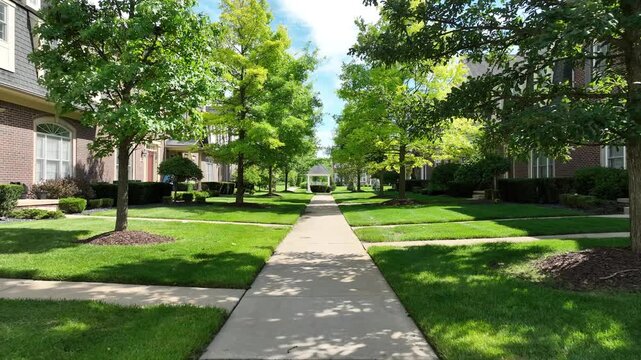 Serene 4K walk-through of a tree-lined condo walkway with brick townhomes, manicured lawns, blue sky, and gazebo&mdash;peaceful suburban community vibe.