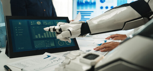 A robot collaborates with a human team in a modern office setting, analyzing data on a touchscreen display, showcasing technology's role in business productivity.Noogenesis
