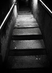 Dark, narrow stairway descends into gloom. Metal handrails flank the stone steps
