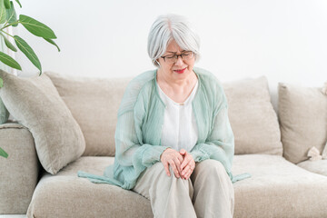 A gray-haired senior woman sitting on a sofa and suffering from knee pain (knee, knee pain)
