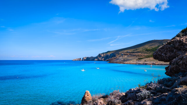 Mediterranean coastal landscape with turquoise sea, rocky shoreline, and distant green mountains under clear blue sky. Natural seaside scenery ideal for travel, nature, and summer design themes.