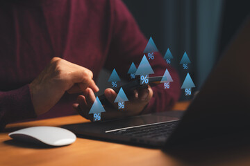 Person using smartphone and laptop with floating upward arrows and percentage symbols, representing financial growth, increasing interest rates, investment returns, economic rise, and digital finance.