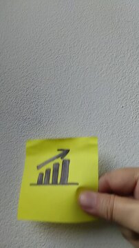 Una toma de cerca de una nota adhesiva de color amarillo con un dibujo de una gr&aacute;fica de barra  ascendente de color negro en un fondo blanco en relaci&oacute;n a las finanzas. Video vertical
