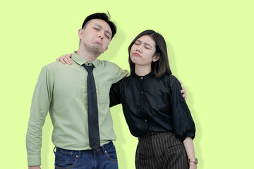 A young Asian couple or colleagues looking exhausted leaning on each other discontented expressions against a plain studio background