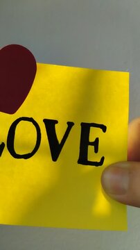 Una toma de cerca de una mano de mujer con una nota adhesiva de color amarillo con la palabra amor y un coraz&oacute;n rojo en movimiento en un fondo blanco. Video vertical