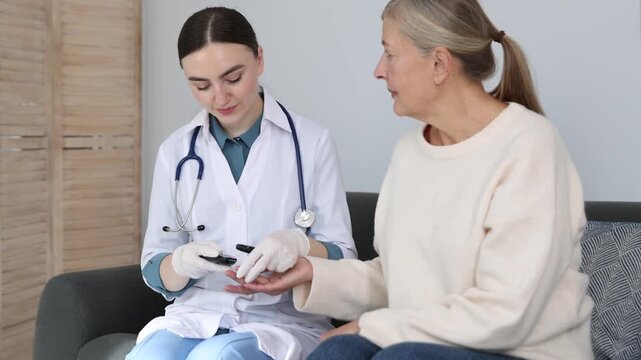 Diabetes. Smiling doctor in medical gloves checking patient's blood sugar level with lancet pen and glucometer on sofa at home. Camera moving towards