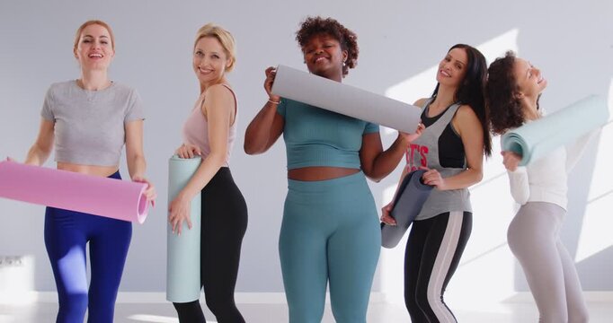 Group of multiracial happy women in sportswear holding yoga mats and dancing together in bright room. Happy female friends having fun during yoga class, hugging and enjoying active healthy lifestyle.