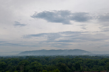 Obraz premium Panoramic landscape of a hazy, majestic volcano mountain dominating the horizon under a dramatic, cloudy morning or evening sky, overlooking a dense rural forest.