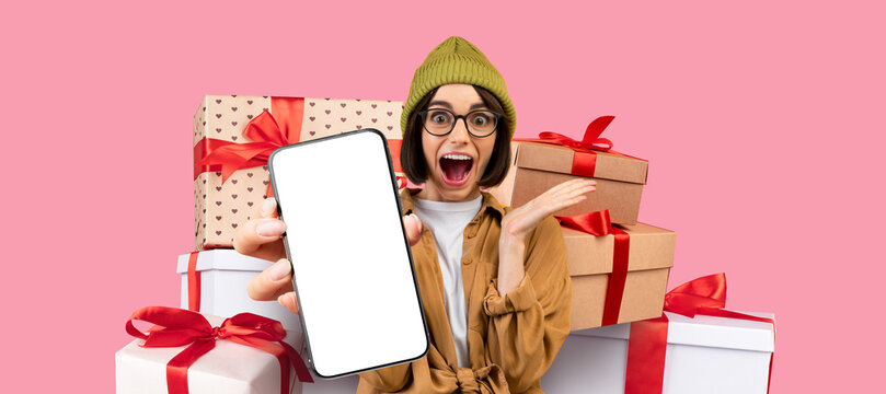 A joyful individual wearing a green beanie and glasses showcases a blank smartphone. Brightly wrapped gifts with red bows create a festive atmosphere. The background adds to the playful mood.