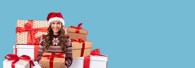 A woman wearing a Santa hat and a cozy sweater smiles while holding a present. Behind her are beautifully wrapped gifts with ribbons, capturing the joyful spirit of holiday celebrations.