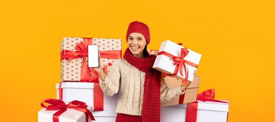 A woman dressed in warm winter attire stands in front of a stack of beautifully wrapped gifts.