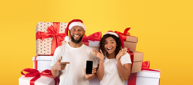 A cheerful couple wearing Santa hats stands in front of a collection of colorful gift boxes. They happily display a smartphone and share an excited moment during the festive season.