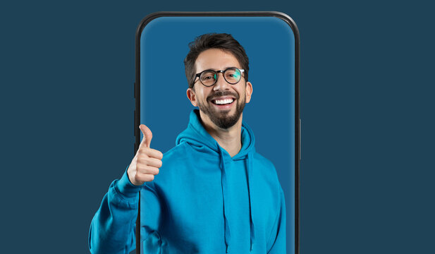 A cheerful man in a blue hoodie gives a thumbs up with a big smile. He appears to be coming out of a smartphone frame against a solid blue background. His expression suggests friendliness.