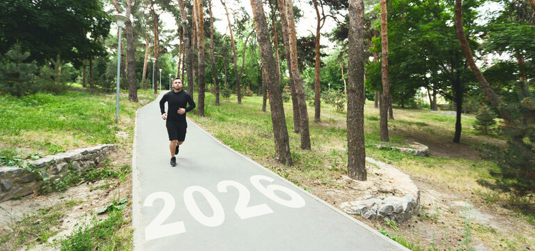 A man jogs along a paved trail in a lush park, surrounded by trees. The year 2026 is painted on the path, indicating a special event or milestone.