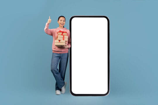 Beautiful young Korean woman stands next to a giant smartphone, holding gift boxes and giving a thumbs up. The studio background is blue, highlighting the mobile app for online shopping. - Powered by Adobe