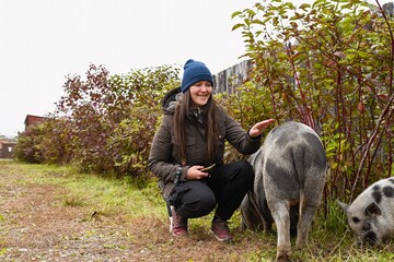 Smiling Russian woman farmer crouching near mini pig on a farm. Warm rural moment showing care and connection between human and animal outdoors.