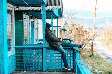 Russian woman sitting on the blue wooden porch of an old rural house in a village in Russia. Autumn day, calm countryside atmosphere and rustic style.