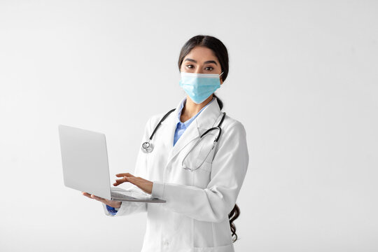 A young Hindu lady doctor in a white coat and protective mask is focused on her laptop in a bright studio. She represents the new normal of remote consultations during social distancing.