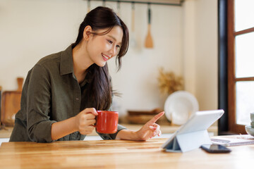 Asian woman drink a cup of coffee and reading news on digital tablet in a kitchen using laptop at home, Activity lifestyles concept